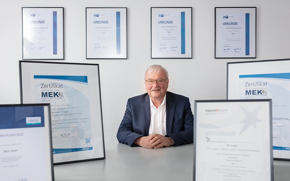 Heinz-Friedrich Kammen, Unternehmensgründer, umgeben von Urkunden und Auszeichnungen, die seine erfolgreiche Ausbildung und Karriere symbolisieren.