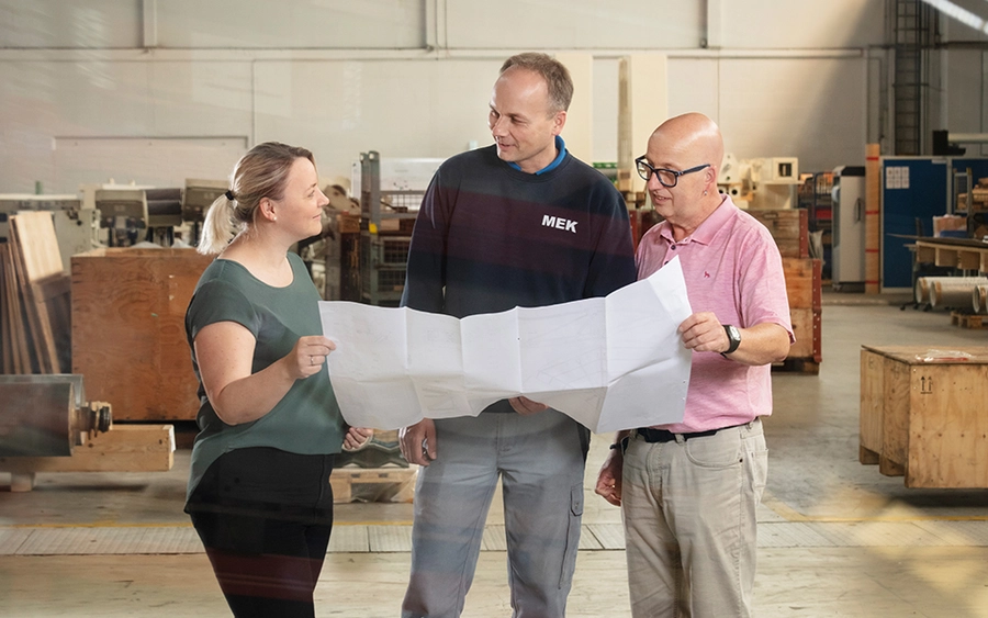 Drei Mitarbeiter in einer Werkshalle betrachten einen großen Faltplan (Zeichnung) und koordinieren sich bei der Planung von technischen Aufgaben.